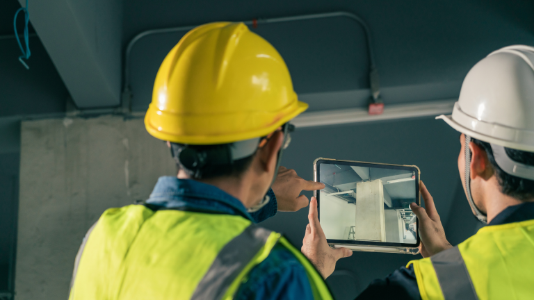 Two engineers in hard hats reviewing building assessment data on a tablet at a mechanical site